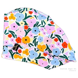 Könnte beinhalten: Eine OP-Haube mit floralem Muster mit weißem Hintergrund und bunten Blumen in Rosa-, Orange-, Gelb- und Blautönen. Die Kappe hat ein schwarzes Etikett mit dem Text "Custom Caps Co."