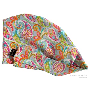Può includere: Una cuffia chirurgica con un vivace motivo paisley nei toni del rosa acceso, verde lime, arancione brillante e turchese su sfondo grigio chiaro. Un'etichetta in tessuto nero con il testo bianco Custom Caps Co. è fissata sul lato della cuffia.