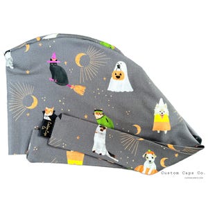 May include: Gray Halloween-themed scrub cap with a matching headband. The cap features illustrations of cats, dogs, and ghosts in costumes, along with moons and stars. The text "Custom Caps Co." is visible on the cap.