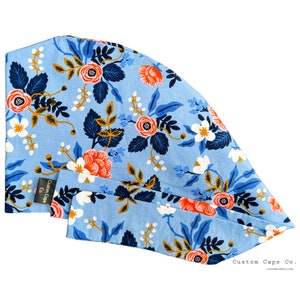 May include: A light blue scrub cap with a floral print featuring white, orange, and blue flowers. The cap has a tie at the back and the text "Custom Caps Co." is printed on the bottom right corner.