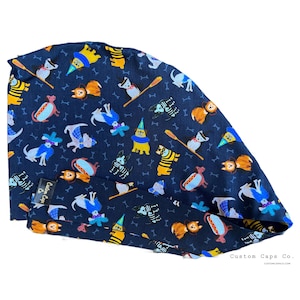 May include: A dark blue scrub cap featuring a repeating pattern of dogs in various costumes, including hot dogs, bumblebees, and party hats, interspersed with small white bones. A black fabric tag reads Custom Caps Co.