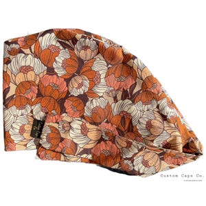 Pode incluir: Um gorro de esfrega com estampa floral marrom, laranja e branco com o texto "Custom Caps Co." e o site "customcapsco.com".