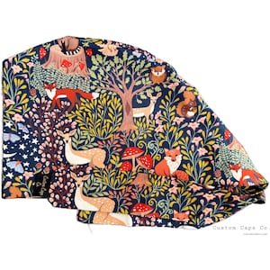 May include: A blue scrub hat with a woodland print featuring deer, foxes, mushrooms, and a large tree. The fabric has a whimsical, colorful design.