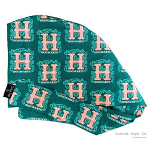 May include: A green fabric headband with a repeating pattern of the Hogwarts crest in a peach colour. The crest features the letter "H" surrounded by a decorative design. The headband is folded in half and the text "Custom Caps Co." is visible in the bottom right corner.