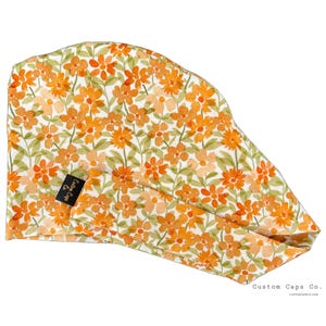 May include: A floral patterned scrub cap featuring orange flowers and green leaves on a white background. The cap has a black tag that says "Custom Caps Co."