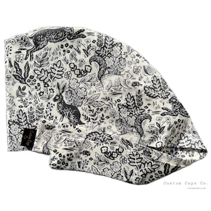 May include: A white and black patterned scrub cap featuring a woodland scene with rabbits, squirrels, and foliage. The cap has a tag that says "Custom Caps Co." and the company's website address.