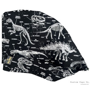 May include: Black surgical cap featuring a white dinosaur skeleton print with labeled bone structures. The cap includes a matching band and the text "Custom Caps Co."