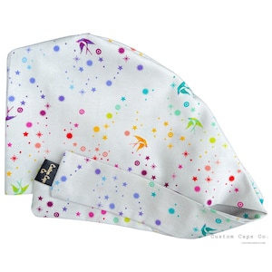 May include: White scrub cap featuring a pattern of colourful stars and swallows in shades of red, orange, yellow, green, blue, and purple. The cap has a matching headband and a black label that says "Custom Caps Co."