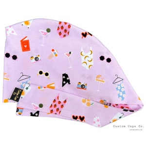May include: A light purple surgical scrub cap with a summer-themed pattern. The pattern includes sunglasses, cameras, cocktails, swimsuits, and strawberries. The cap has a tag that says "Custom Caps Co."