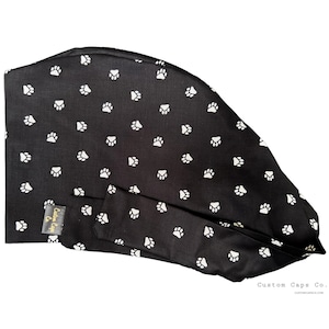 Könnte beinhalten: Eine schwarze OP-Haube mit einem Muster aus weißen Pfotenabdrücken. Die Haube hat einen Bindebandverschluss und ein kleines Etikett mit der Aufschrift "Custom Caps Co.". Die Worte "Custom Caps Co." und eine Webadresse sind unten rechts aufgedruckt.