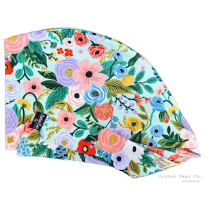 May include: A turquoise blue scrub cap with a floral print featuring pink, purple, and yellow flowers. The cap has a label that says "Custom Caps Co."