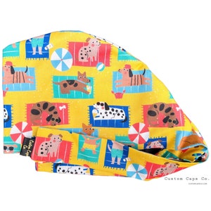 May include: A yellow surgical scrub cap with a repeating pattern of cartoon dogs relaxing on beach towels. The dogs are depicted in various poses, some wearing sunglasses or drinking beverages. The cap also features beach umbrellas and the text "Custom Caps Co."