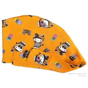 May include: An orange scrub cap featuring a repeating pattern of cartoon cats wearing striped shirts and bows, along with small gift boxes. A black fabric tag with the white text Custom Caps Co. is attached to the side of the head covering.