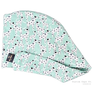 May include: A mint green surgical scrub cap featuring a repeating pattern of white and grey dalmatian dogs. The cap has a black label that reads "Custom Caps Co." and the words "CustomCapsCo.com" are printed on the bottom right.