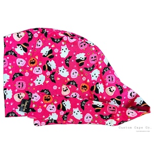 May include: A bright pink scrub cap featuring a repeating pattern of white ghosts, black bats, and orange jack-o'-lanterns. The fabric includes small white dots and a black tag with the text Custom Caps Co. sewn onto the side of the headband.