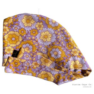 Puede incluir: Un gorro de quirófano de tela con estampado floral morado y amarillo con una etiqueta marrón que dice "Custom Caps Co."