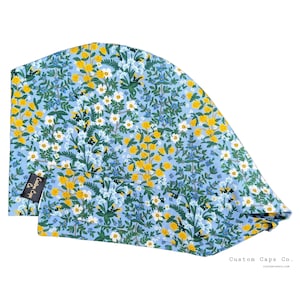 May include: A light blue surgical scrub cap with a floral pattern. The cap features a repeating design of yellow, white, and blue flowers with green foliage. The text "Custom Caps Co." is visible on a small tag and on the bottom right.