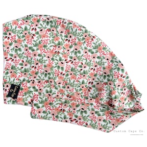 Puede incluir: Un gorro quirúrgico con estampado floral, con fondo blanco adornado con rosas color melocotón, bayas rojas y hojas verdes. El gorro tiene una etiqueta negra con texto blanco: "Custom Caps Co."
