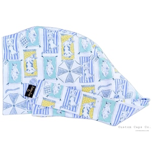 May include: A white surgical scrub cap featuring a repeating pattern of cartoon bears on beach towels, umbrellas, and beach bags. The design includes blue and yellow accents. The cap has a small black tag with white text.