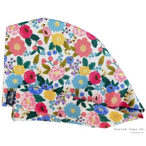 Peut inclure: Une toque chirurgicale blanche ornée d'un motif floral vif. Le motif présente des fleurs roses, jaunes et bleues avec des feuilles vertes et des baies bleues. Le bonnet porte le texte "Custom Caps Co." sur le côté.