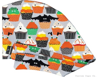 May include: A light gray surgical scrub cap and matching face mask featuring a Halloween-themed cupcake pattern. The cupcakes have orange, yellow, green, and black frosting, with bat and spider decorations. The brand name "Custom Caps Co." is visible.