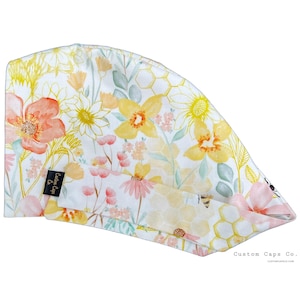 May include: A white scrub cap with a floral print featuring yellow, pink, and green flowers. The fabric has a honeycomb pattern with bees. The text "Custom Caps Co. customcapsco.com" is printed on the bottom right corner.