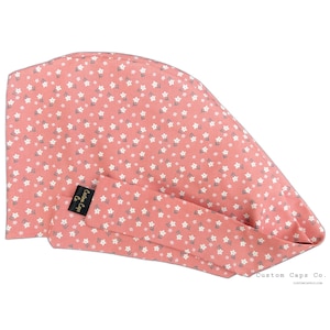 May include: A pink surgical cap with a white floral pattern. The cap has a tie at the back and a small label that says "Custom Caps Co."
