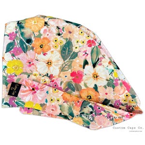 May include: A floral print scrub cap with a watercolour design. The cap features pink, orange, yellow, and white flowers and green leaves. A black tag reads "Custom Caps Co."