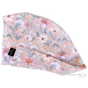 May include: Peach-colored scrub cap with a floral pattern of white and pink flowers and gray leaves. The cap has a matching headband. The label reads "Custom Caps Co."