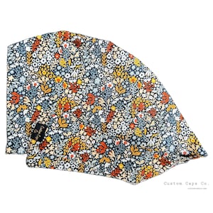 Pode incluir: Uma touca cirúrgica com padrão floral. A touca tem um fundo azul marinho escuro com uma variedade colorida de flores e folhas em tons de amarelo, laranja, branco e azul. A etiqueta diz "Custom Caps Co."