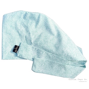 Può includere: Un berretto chirurgico blu chiaro con un motivo a pois bianchi. Il berretto ha un laccio sul retro e una piccola etichetta nera che dice "Custom Caps Co."
