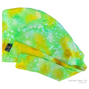 May include: A lime green and yellow surgical scrub cap with a watercolor-like pattern. The cap has a tie-back design and a small black tag that reads "Custom Caps Co."
