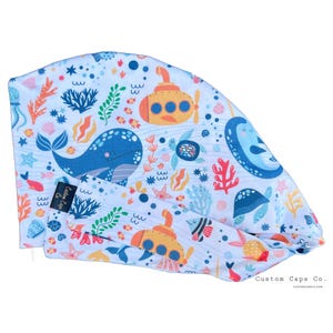 May include: A blue and white surgical cap with a colorful underwater scene featuring a whale, a submarine, and sea life. The cap has a tie at the back and the text "Custom Caps Co. CUSTOMCAPSCO.COM" is printed on the bottom right corner.