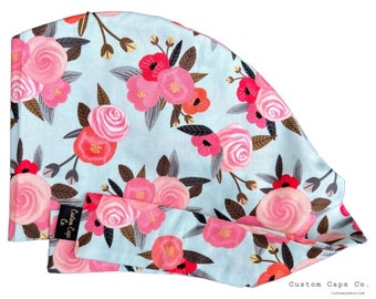Puede incluir: Una cofia quirúrgica azul claro con un estampado floral con rosas rosas y rojas. La cofia tiene un lazo en la parte posterior y el texto "Custom Caps Co. CUSTOMCAPSCO.COM" está impreso en la esquina inferior derecha.