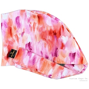 May include: A patterned scrub cap in pink, orange, and purple hues, with a matching headband. The design resembles brushstrokes. A small black label reads "Custom Caps Co."