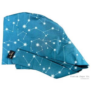 Op de afbeelding: Teal chirurgische scrubcap met een wit sterrenbeeldpatroon. De cap heeft een lange stropdas en een klein zwart label met de tekst "Custom Caps Co.". De woorden "Custom Caps Co." staan ook rechtsonder gedrukt.