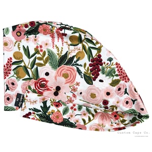 May include: A white scrub cap with a floral print featuring pink, red, and green flowers. The cap has a black label with the text "Custom Caps Co."