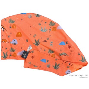 May include: A bright orange scrub cap with a colorful desert-themed print featuring cacti, lizards, and other desert flora and fauna. The cap has a label that reads "Custom Caps Co."