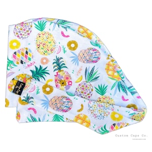 May include: White scrub cap featuring a vibrant pineapple design. The pattern includes yellow, pink, green, and blue pineapples, with floral and geometric details. The cap has a black label that says "Custom Caps Co."