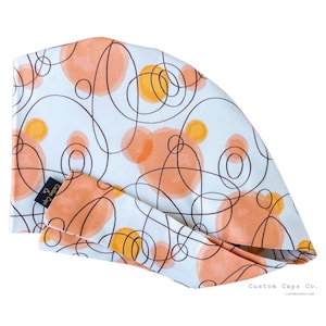 May include: A white scrub cap featuring a pattern of overlapping dark brown circular line drawings and soft peach and yellow watercolour-style circles. A small black fabric tag on the side displays the text Custom Caps Co.