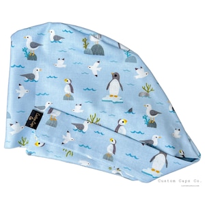 May include: Light blue surgical scrub cap with a repeating pattern of seagulls, penguins, and ocean waves. The cap is made of a soft fabric and features a coordinating band. The text "Custom Caps Co." is printed on the cap.