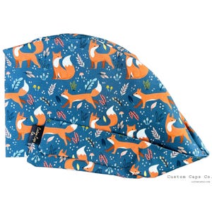 May include: A blue surgical scrub cap with a repeating pattern of orange foxes, white-tipped tails, and botanical elements. The cap is made of a soft fabric and features a tie-back design. The text "Custom Caps Co." is visible.