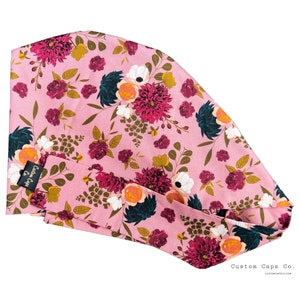 Pode incluir: Uma touca cirúrgica floral rosa com um padrão repetido de flores bordô, brancas e amarelas e folhas verdes. A touca tem uma gravata combinando e uma pequena etiqueta preta que diz "Custom Caps Co."