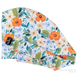 Pode incluir: Uma touca cirúrgica com estampa floral com papoulas laranja, flores azuis e creme e folhas verdes em um fundo branco. A touca tem uma gravata nas costas e uma pequena etiqueta preta que diz "Custom Caps Co."