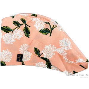 May include: Peach-colored surgical scrub cap with a white floral pattern. The cap has a tie at the back and a small label that says "Custom Caps Co."
