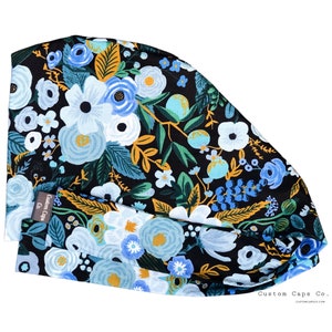 May include: A black scrub cap with a floral pattern featuring blue, white, and yellow flowers on a dark green background. The cap has a label that reads "Custom Caps Co."