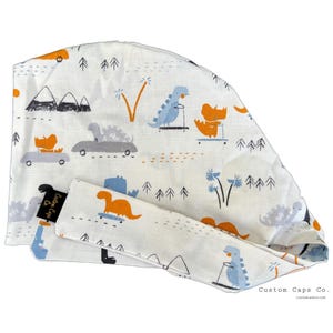 May include: White surgical scrub cap and matching headband featuring a playful dinosaur print. The design includes orange, blue, and gray dinosaurs on skateboards and in cars, set against a backdrop of trees and mountains.