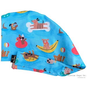 May include: A blue surgical cap with a pattern of dogs enjoying summer activities like swimming, floating on a banana, and playing with a beach ball. The text "Custom Caps Co." is printed on the bottom right corner of the cap.