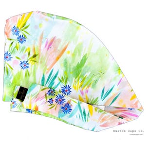May include: A white scrub cap with a colorful floral print featuring blue flowers, green leaves, and pink and yellow accents. The cap has a black label with the text "Custom Caps Co."