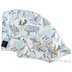 Puede incluir: Una gorra quirúrgica blanca con un patrón repetido de animales árticos, incluyendo zorros, búhos y renos, con acentos dorados y negros. La gorra tiene un fondo azul claro y verde con árboles estilizados. El logotipo de Custom Caps Co. es visible.